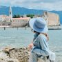 A woman in a blue hat gazes at the sea, with a castle in the background, embodying the spirit of travel and exploration.