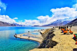 A group of travelers stands on a serene lake shore, with majestic mountains rising in the background, enjoying nature's beauty